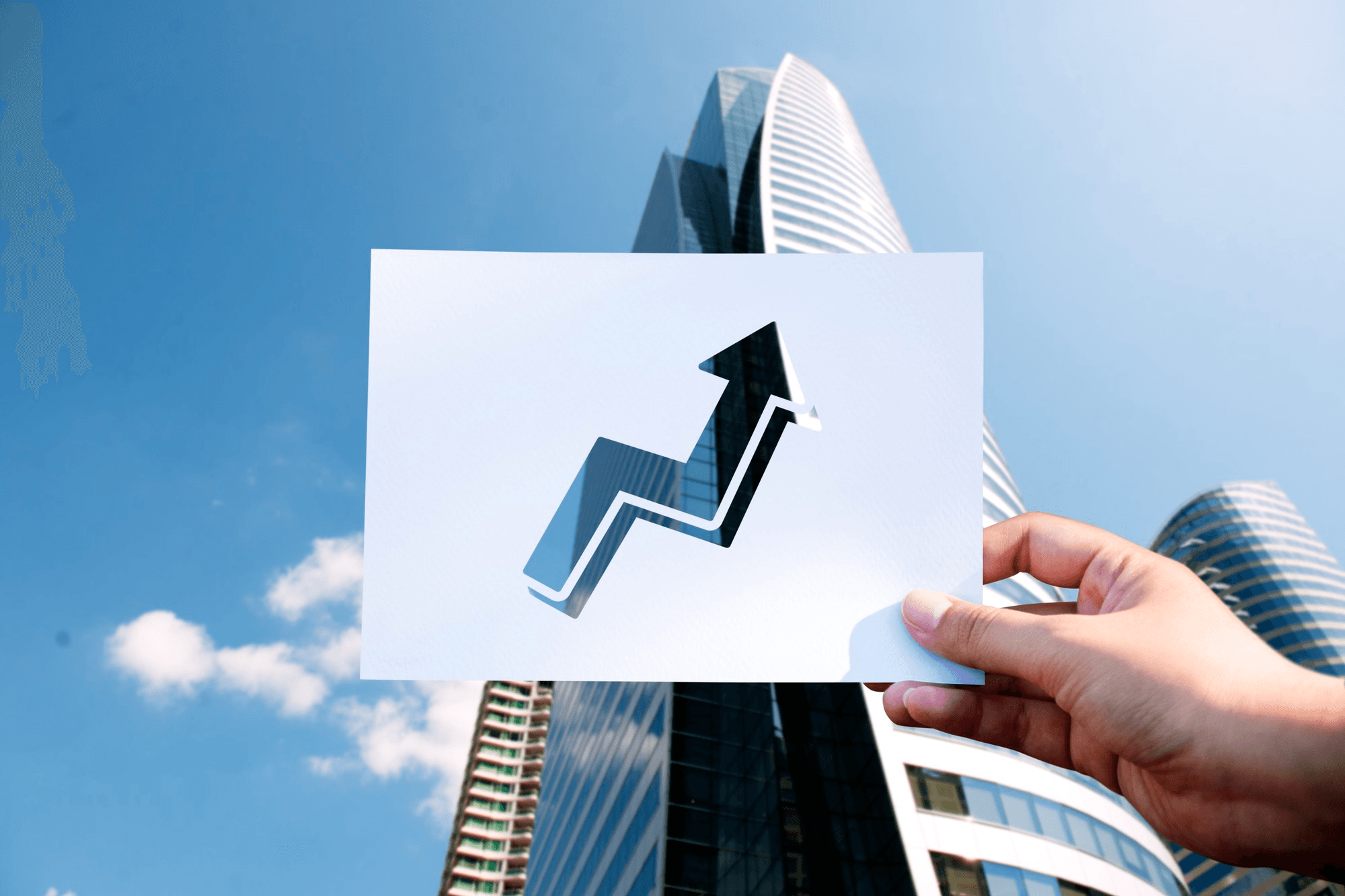 A hand holding a cut-out of an upward trending arrow in front of a modern skyscraper to show the role of real estate risk management software